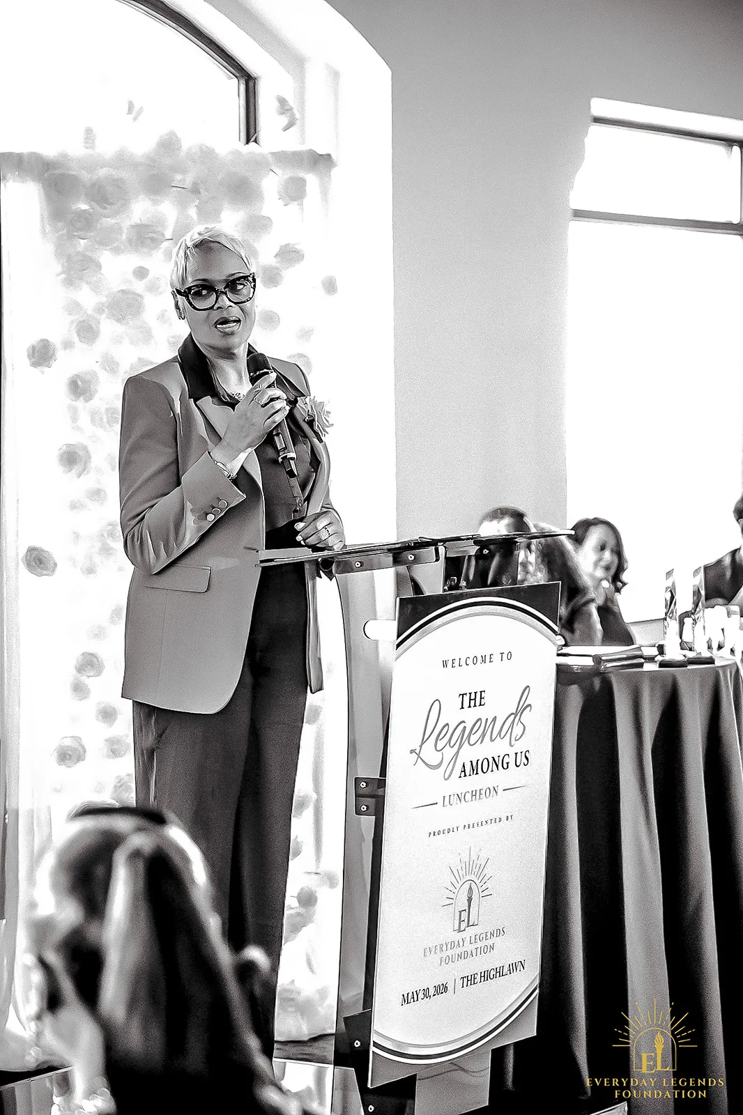 A speaker addresses the room from the lectern at the Legends Among Us luncheon at The Highlawn, beside the welcome sign.