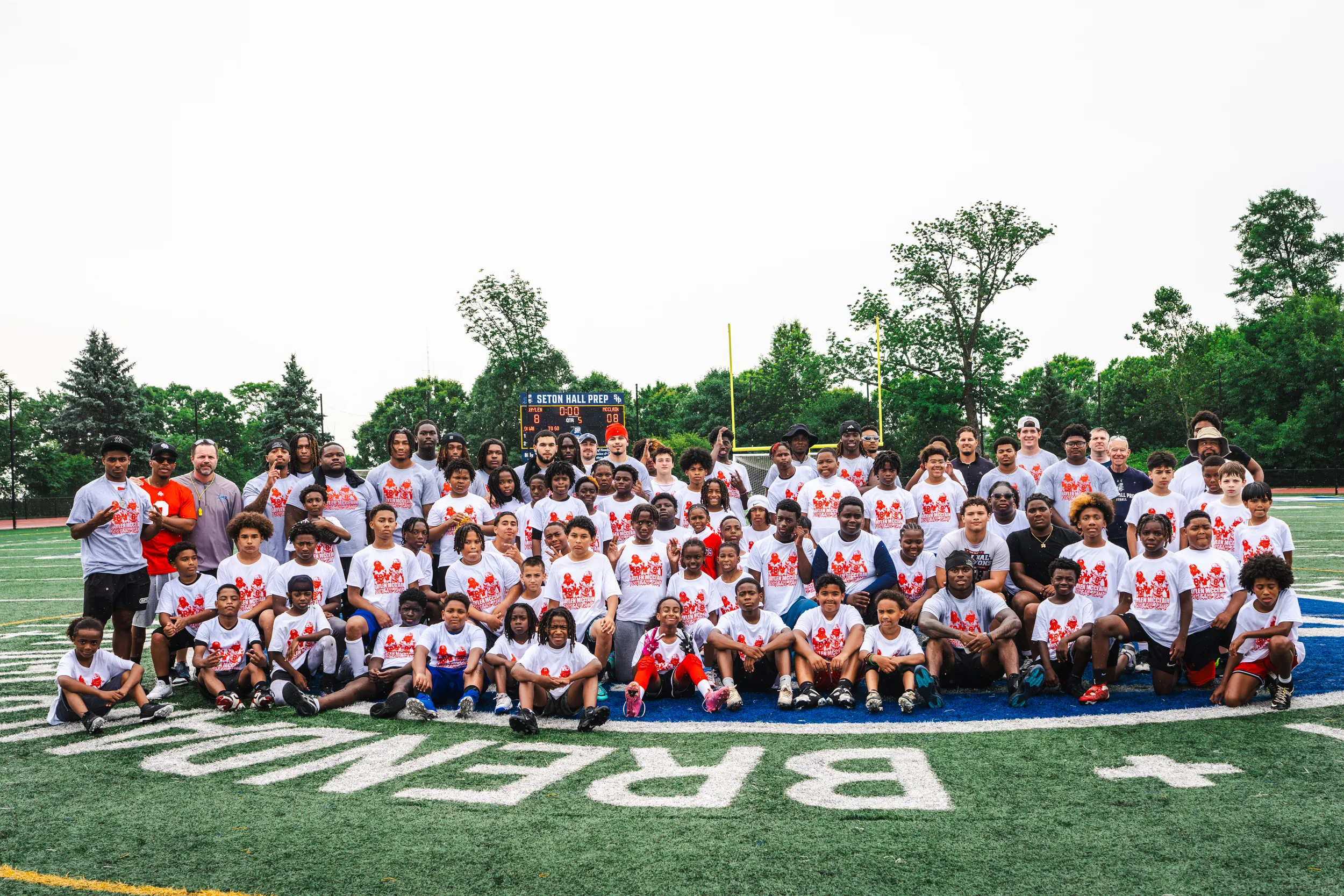 Campers, coaches, and volunteers in camp T-shirts pose together on the Seton Hall Prep football field.